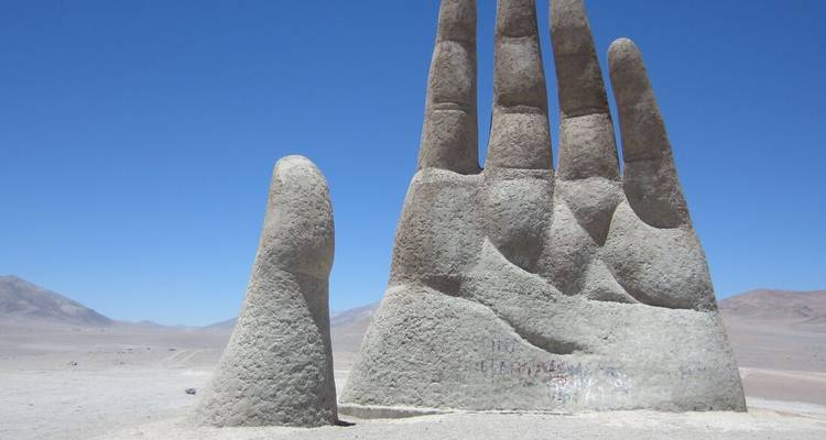 Massief betonnen sculptuur van een hand die oprijst uit een lege woestijnvlakte onder een heldere blauwe hemel.