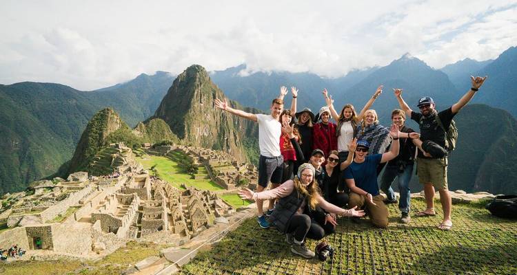 Vrolijke groep reizigers steekt armen omhoog op een terras met uitzicht op de ruïnes van Machu Picchu.