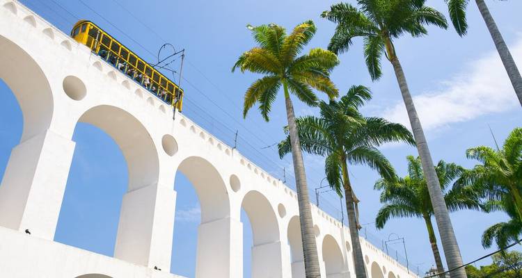 White arches of Lapa Aqueduct with yellow tram passing, framed by tall palm trees and blue sky.