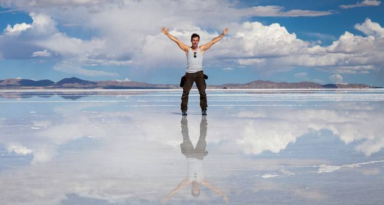 Voyageur debout les bras levés sur la surface réfléchissante du Salar d'Uyuni sous des nuages dramatiques