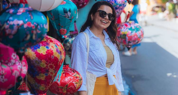 Smiling woman in stylish outfit stands beside rows of colourful lanterns lining a historic street.