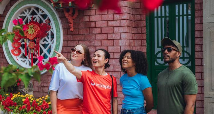 Guide points out decorations on a festive building façade while diverse travellers look on amid red bougainvillea blooms.