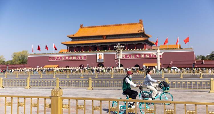 Radfahrer fahren an dem mit roten Fahnen geschmückten Tiananmen-Tor vorbei, während sich Menschenmengen auf dem weiten Platz hinter dem goldgedeckten Wahrzeichen versammeln.