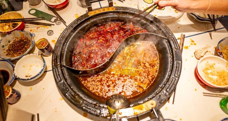 Una olla caliente humeante de yin-yang llena de caldo rojo picante y caldo dorado más suave se encuentra en el centro de una mesa compartida animada.