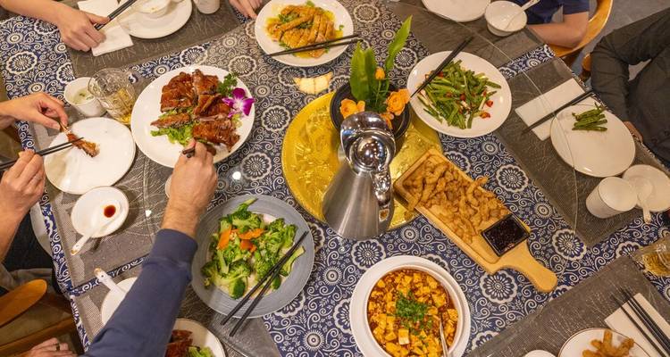Vista aérea de una mesa de banquete donde los comensales comparten múltiples platos chinos, vegetales vibrantes y tofu sobre un mantel estampado.