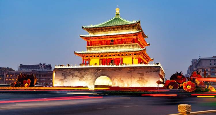 La Torre de la Campana de Xi'an brillando con luces cálidas contra un cielo crepuscular y el tráfico de la ciudad en movimiento.