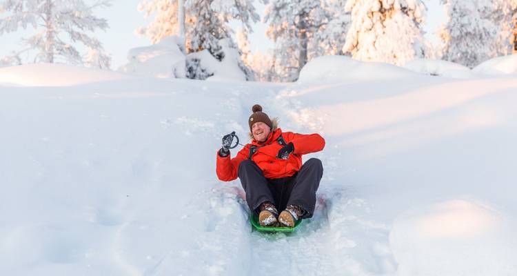 Smiling sledder sliding down a snowy hill surrounded by frost-covered trees on a sunny winter day.