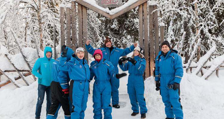 Cheerful group in blue thermal suits celebrating at the entrance of a snowy husky kennel.