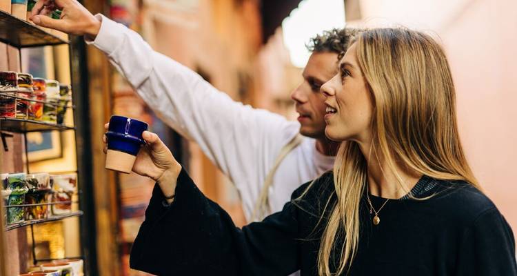 Een stel verkent een smalle souk in Marrakech en bekijkt kleurrijke keramische kopjes die aan een stallenwand hangen.