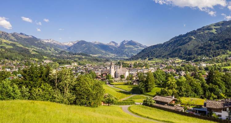 Ciudad del valle verde de Kitzbühel enclavada entre picos alpinos con chalets dispersos y campanario de iglesia visible bajo un cielo soleado.