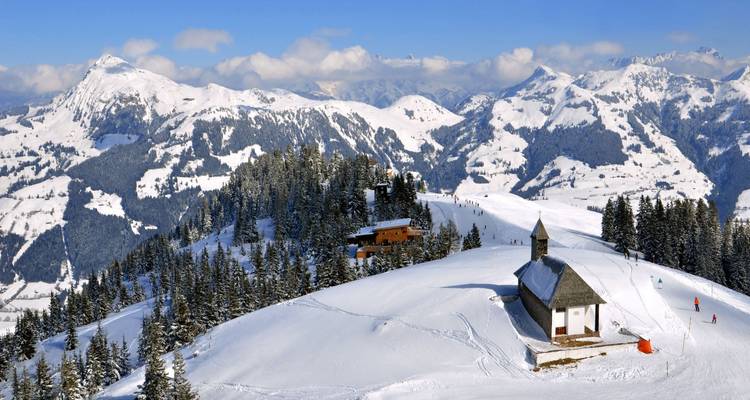 Cresta alpina cubierta de nieve con una pequeña capilla y esquiadores disfrutando de pistas invernales prístinas contra un telón de fondo de picos escarpados.