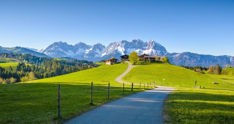 Sereno camino rural que conduce a una granja en la cima de una colina con las montañas Wilder Kaiser cubiertas de nieve al fondo bajo cielos azules despejados.