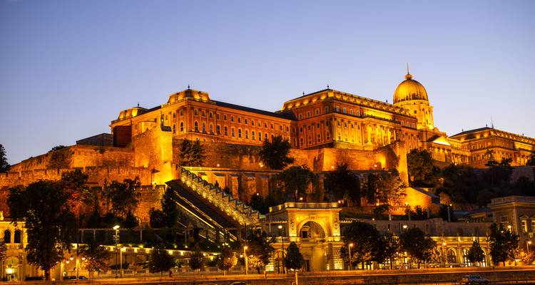 Castillo de Buda iluminado y murallas circundantes brillando doradas al crepúsculo sobre el Danubio en Budapest.