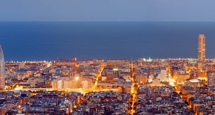 Vista nocturna elevada del paisaje urbano cuadriculado de Barcelona iluminado contra el horizonte del Mar Mediterráneo.