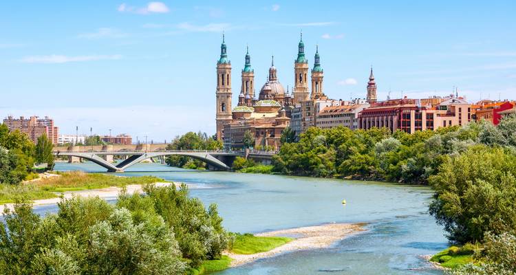 Vista panorámica de la Basílica del Pilar de Zaragoza junto al tranquilo río Ebro bajo cielos azules despejados.