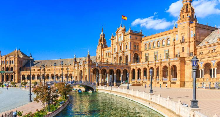 Los arcos ornamentados y las torres de la Plaza de España reflejados en su canal bajo cielos azul vívido.