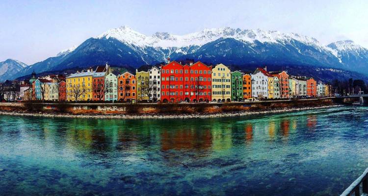 Casas de colores en fila a lo largo del río Inn con los Alpes nevados elevándose detrás y sus reflejos espejados en el agua.