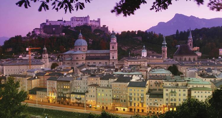 Twilight view over Salzburg with the historic fortress and cathedral above pastel-lit rooftops, mountains silhouetted behind.