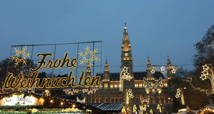 Festive lights spell out "Frohe Weihnachten" in front of an illuminated neo-gothic city hall at a Christmas market after dusk.