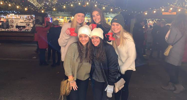 Five young women bundled in winter clothes pose with festive mugs under string lights at a Christmas market.