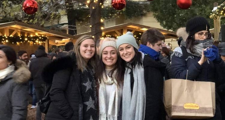 Three smiling friends in winter attire stand close together at a busy outdoor Christmas market.