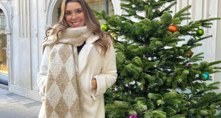 A young woman in a cozy coat and scarf stands beside a decorated evergreen on a city street.