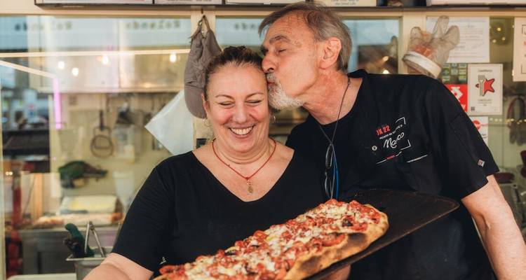 Ein lokaler Koch küsst eine lächelnde Frau liebevoll, während er ein Tablett mit frisch gebackener römischer Pizza al taglio präsentiert.
