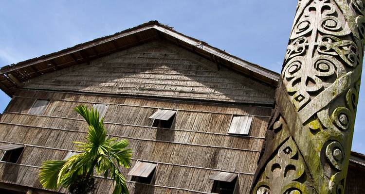 Une maison longue traditionnelle de Bornéo en bois se dresse sous un ciel dégagé à côté d'un mât totémique sculpté.