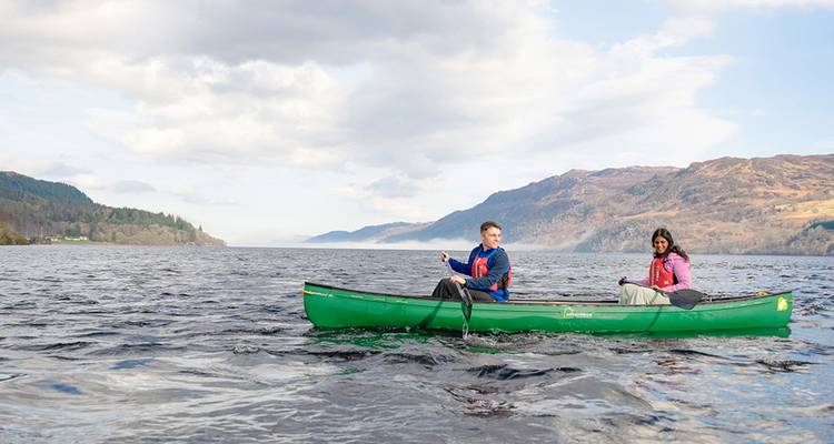 Two paddlers in a green canoe glide across a vast Scottish loch surrounded by rolling hills