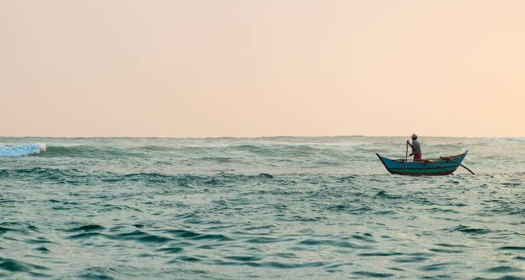 Un pescador solitario se encuentra en una pequeña embarcación de madera sobre suaves mares turquesa al amanecer.