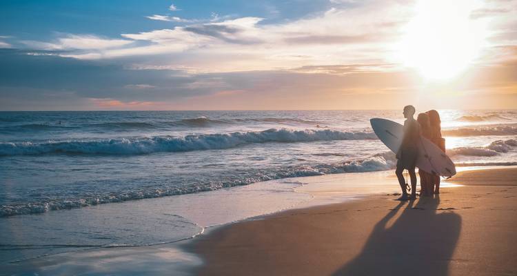 Pareja en silueta con tablas de surf parada sobre arena húmeda en un atardecer dorado con olas rompiendo.