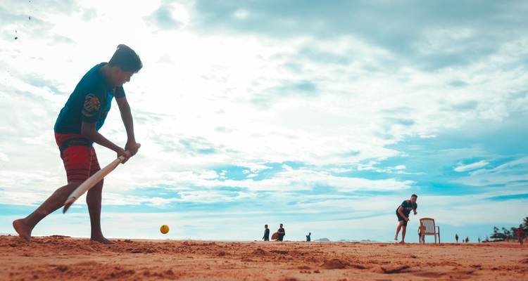 Los niños juegan cricket en la playa bajo un cielo brillante, bateador a media oscilación en la orilla arenosa.