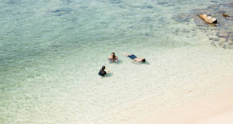 Tres personas se relajan y flotan en aguas marinas cristalinas y poco profundas junto a una orilla arenosa.