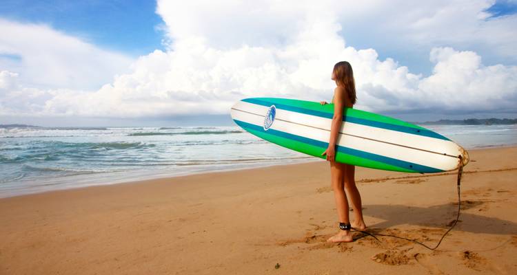 Mujer joven sosteniendo una tabla de surf verde y blanca contempla las olas que rompen en una playa tropical vacía.