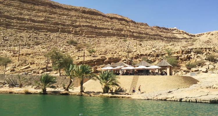Restaurante de terraza de piedra sombreado por sombrillas se encuentra junto a piscinas de color esmeralda y palmeras en un oasis del desierto.
