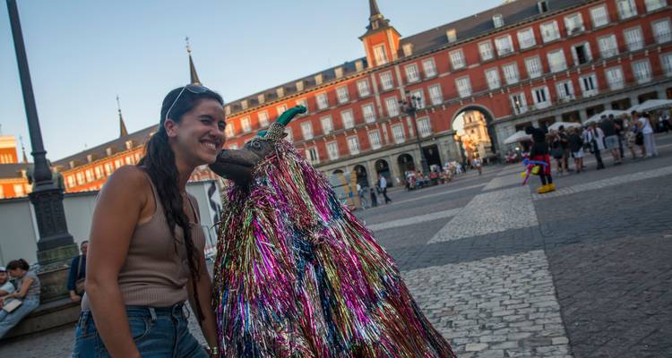 Un visitante sonríe al lado de un artista callejero cubierto de oropel colorido en la Plaza Mayor de Madrid llena de gente al atardecer.