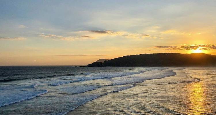 Hermosa puesta de sol dorada sobre olas ondulantes y un acantilado lejano visto desde la orilla.