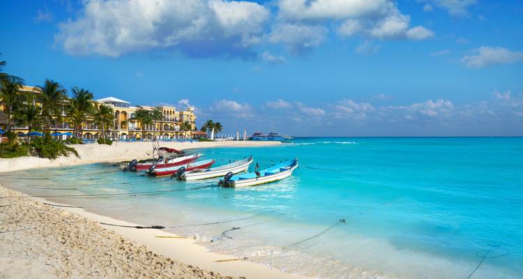 Bunte kleine Boote, die in kristallklarem türkisfarbenem Wasser vor einem weißen Strand vertäut sind, der von Palmen und niedrigen Resortgebäuden gesäumt ist.