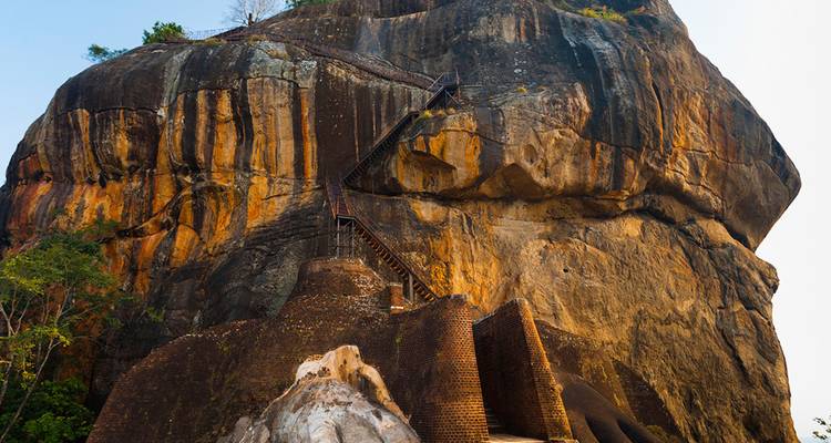 Imposante forteresse rocheuse aux stries orange avec un escalier abrupt montant jusqu'au sommet, entourée de verdure.