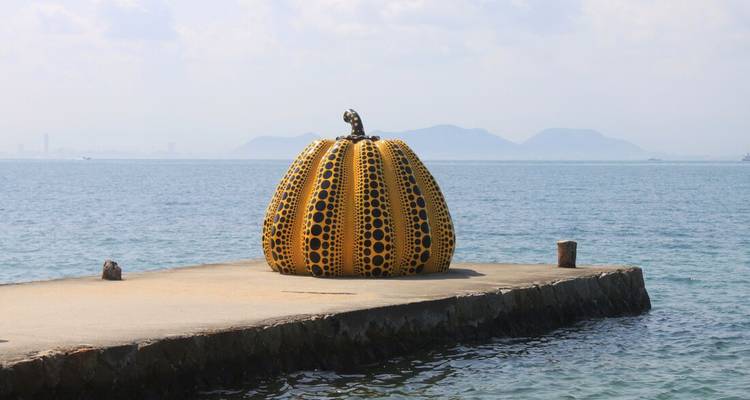 La sculpture de citrouille jaune à pois iconique repose sur une jetée en béton face à une mer bleue calme.