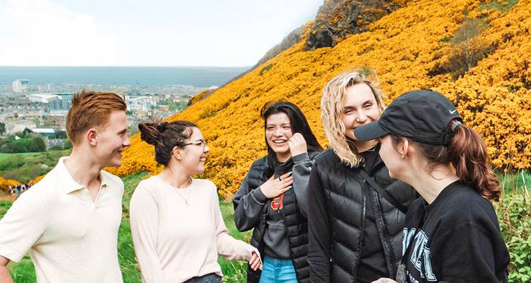Grupo de amigos ríe junto a una ladera cubierta de tojo amarillo con la ciudad y el mar a la distancia