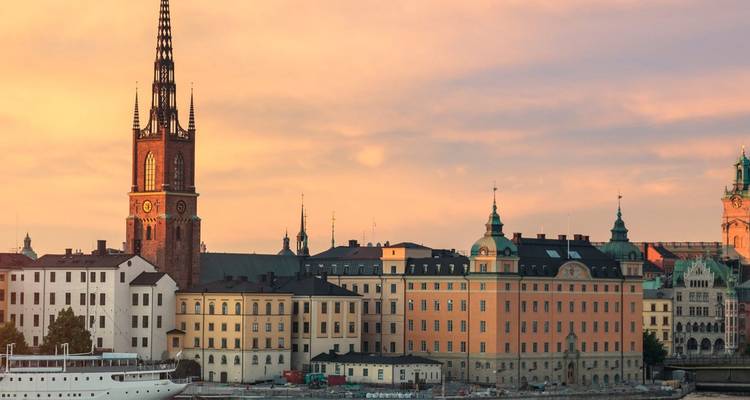 Vista cálida de la hora dorada del paseo marítimo de Estocolmo que muestra la alta aguja de la Iglesia de Riddarholmen y los históricos edificios de colores pastel a lo largo del muelle, con un crucero blanco en aguas tranquilas en primer plano.