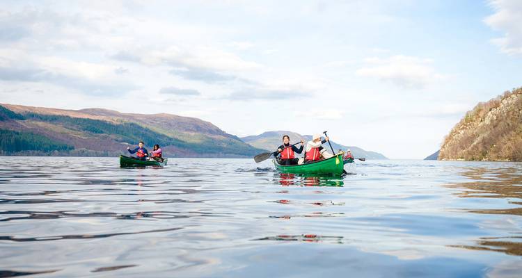 Deux canoës avec des pagayeurs glissent sur un lac calme entouré de collines ondulantes sous un ciel lumineux.