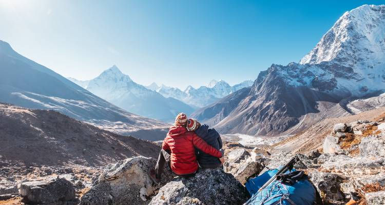 Ein Trekkingpaar in Daunenjacken sitzt auf Felsen und bewundert die schneebedeckte Everest-Gebirgskette.