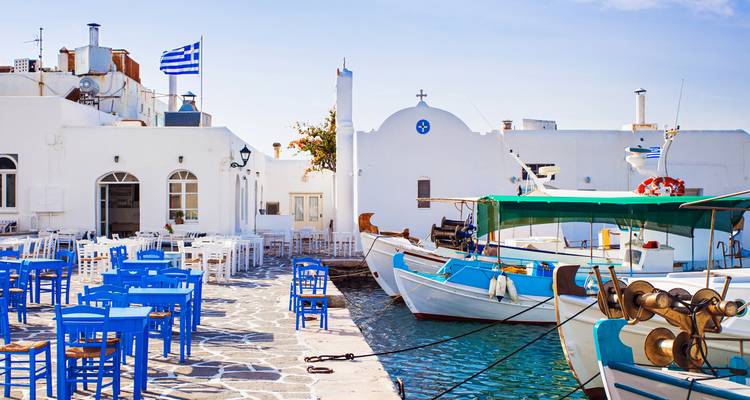 Pintoresco puerto con barcos pesqueros, sillas azules y tabernas encaladas bajo la bandera griega