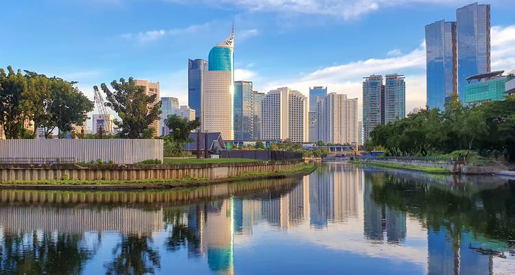 Horizon moderne de Jakarta reflété dans un canal calme avec de la verdure et un ciel bleu au-dessus.
