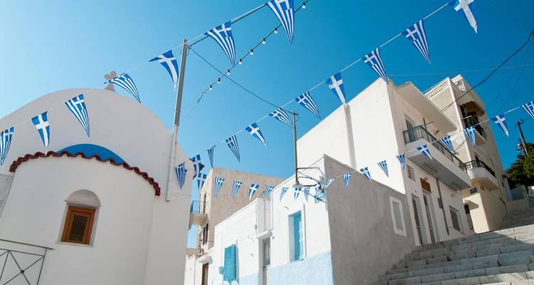 Las banderas griegas azules y blancas ondean sobre casas encaladas y una pequeña capilla bajo un cielo brillante.