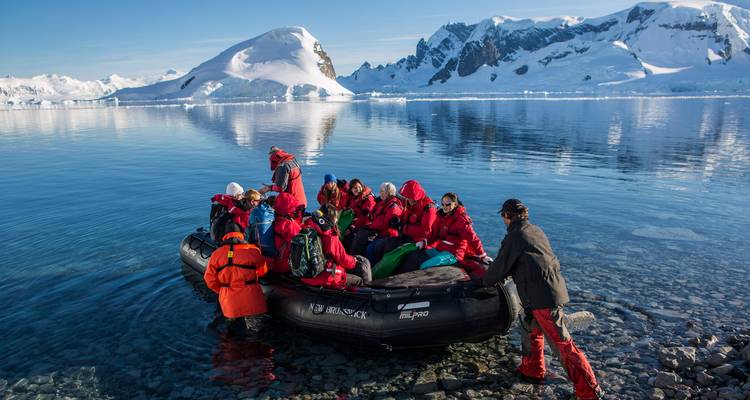 Grupo de viajeros con parkas rojas subiendo a un Zodiac en una bahía antártica clara y cristalina.