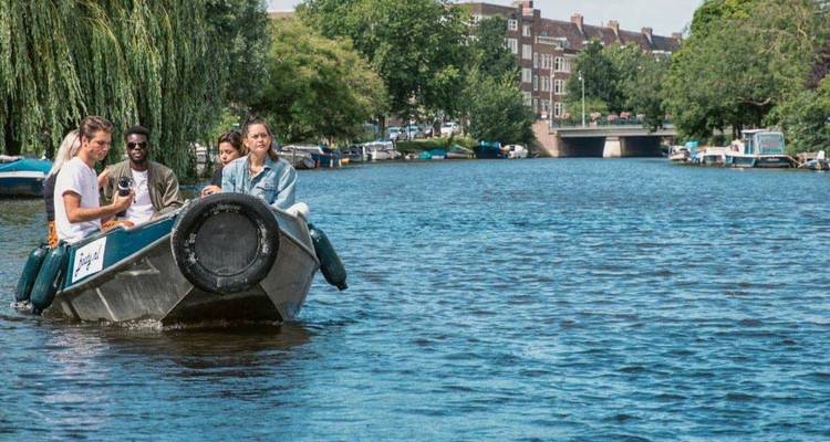 Jonge reizigers varen met een klein motorbootje langs een groene stadskanaal met bakstenen huizen langs het water.