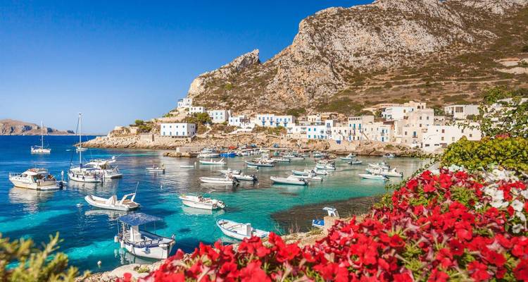 Puerto de aguas turquesas con pequeños botes y casas encaladas enmarcadas por flores rojo brillante y acantilados escarpados.